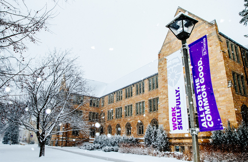 Aquinas Hall is seen through falling snow February 2, 2016. Banners reading "Work Skillfully" and "All for the Common Good" are at right.