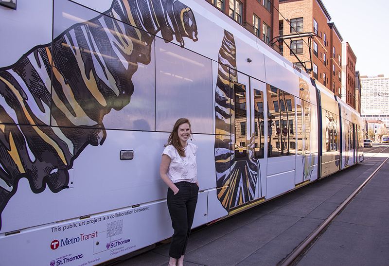 Sarah Nelson poses in front of a light rail car featuring her design for Earth Week. The unique display is the culmination of a partnership between students at the University of St. Thomas, St. Paul-based artist Sarah Nelson and Metro Transit.