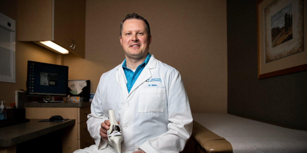 Orthopedic surgeon and University of St. Thomas alumni, Nicholas Weiss ’92, sits for a portrait holding a knee replacement model