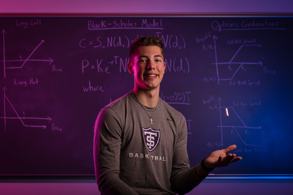 Senior Tommy Anderson, an Actuarial Science major and basketball player, poses for a photo in front of actuarial equations on a blackboard.