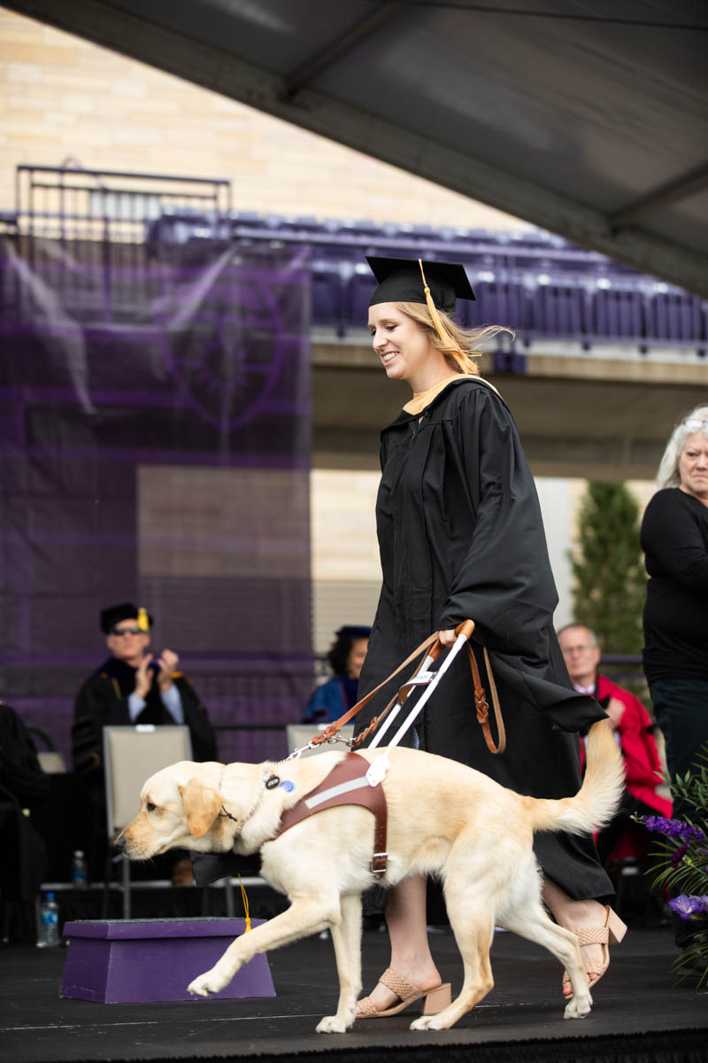 A student walks across stage with an assistance dog during the 2022 Graduate Commencement ceremony for the College of Arts and Sciences, the Morrison Family College of Health and the School of Engineering in St. Paul on May 22, 2022.