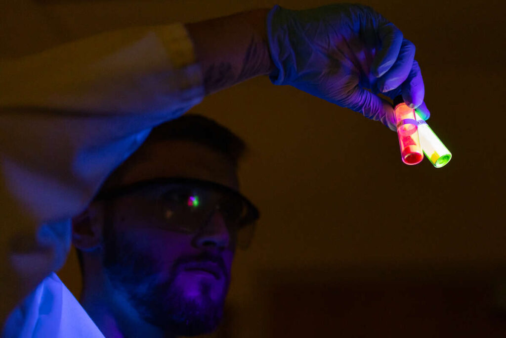 Undergraduate students hold vials up to an ultraviolet light in a lab during the Chemistry Summer Research program on July 26, 2023, in Owens Science Hall in St. Paul.