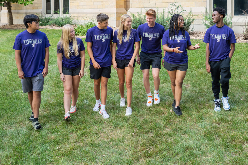 A group of 7 students laugh and talk as they walk across a grass area on the St. Thomas campus