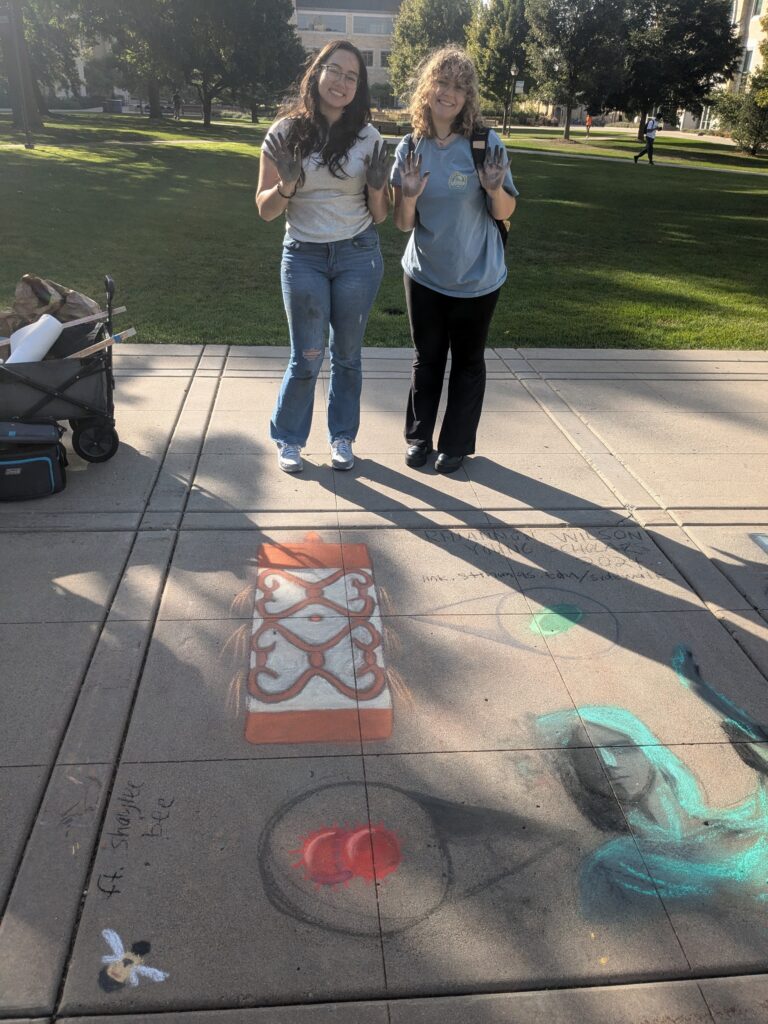 Nayeli and her friend stand, smiling in front of their drawn images holing up their hands which are covered in chalk. 
