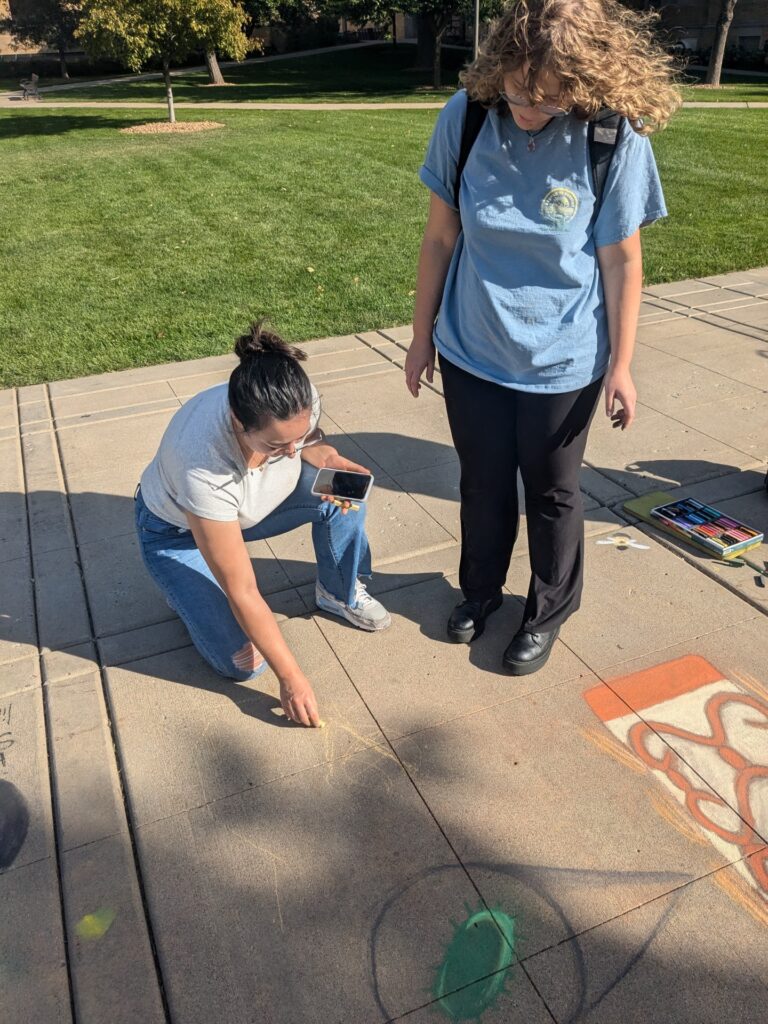 Rhiannon kneels to draw on the sidewalk while her friend stands and watches. 