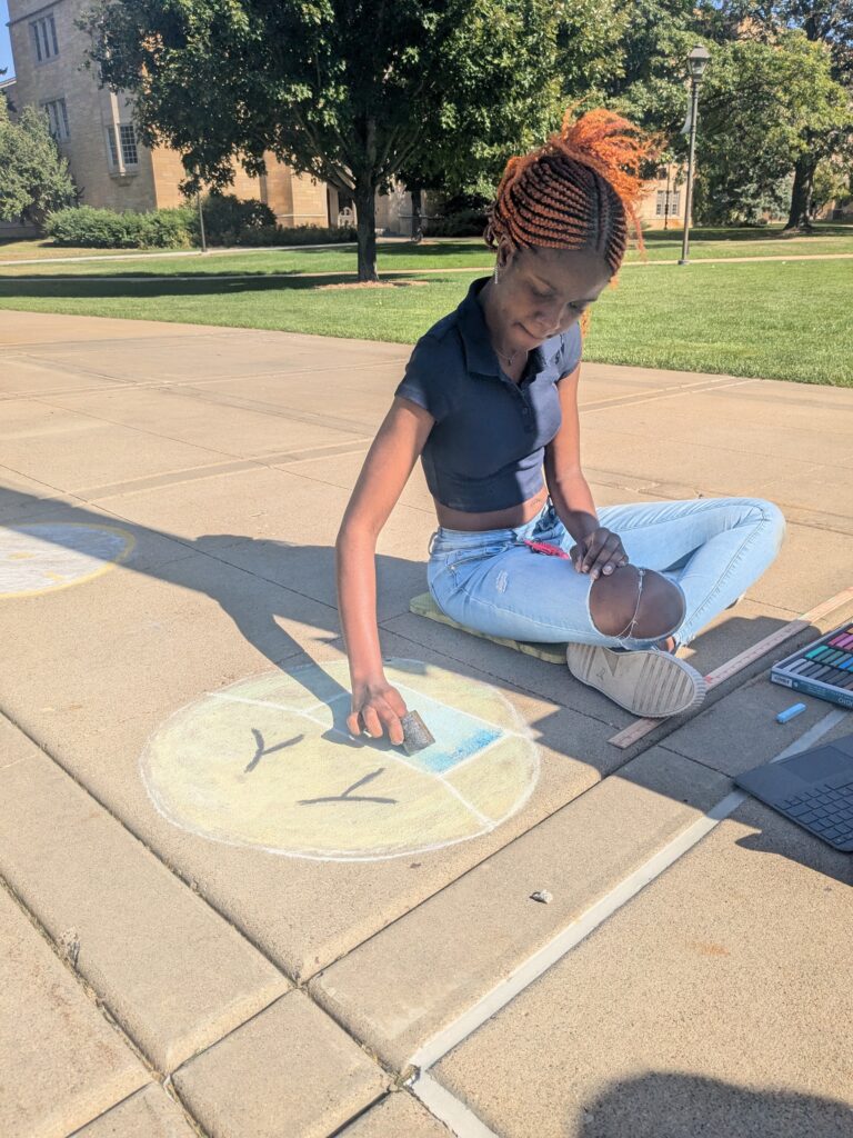 Student Celeste Conteh sits on the ground drawing an image of an emoji face wearing a mask. 