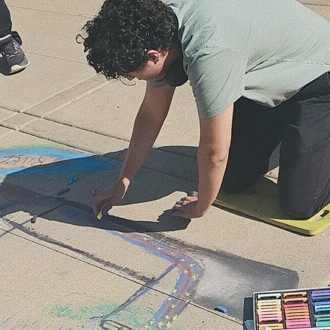 Student Hapaki Lorenzo kneels on the ground drawing with yellow piece of chalk. 
