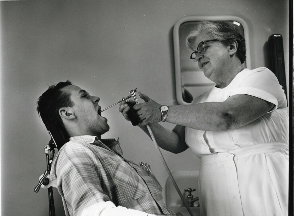 A man opens his mouth as a female nurse examines his throat with a metal tool.