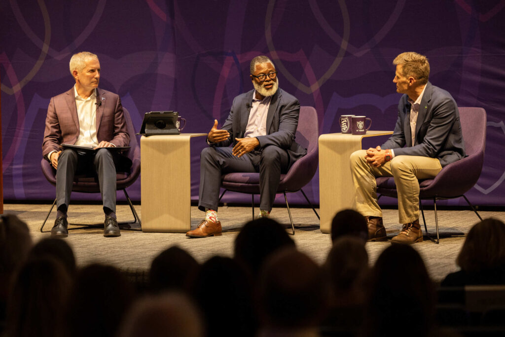 A medium close up shot of three speakers having a discussion in front of an audience.