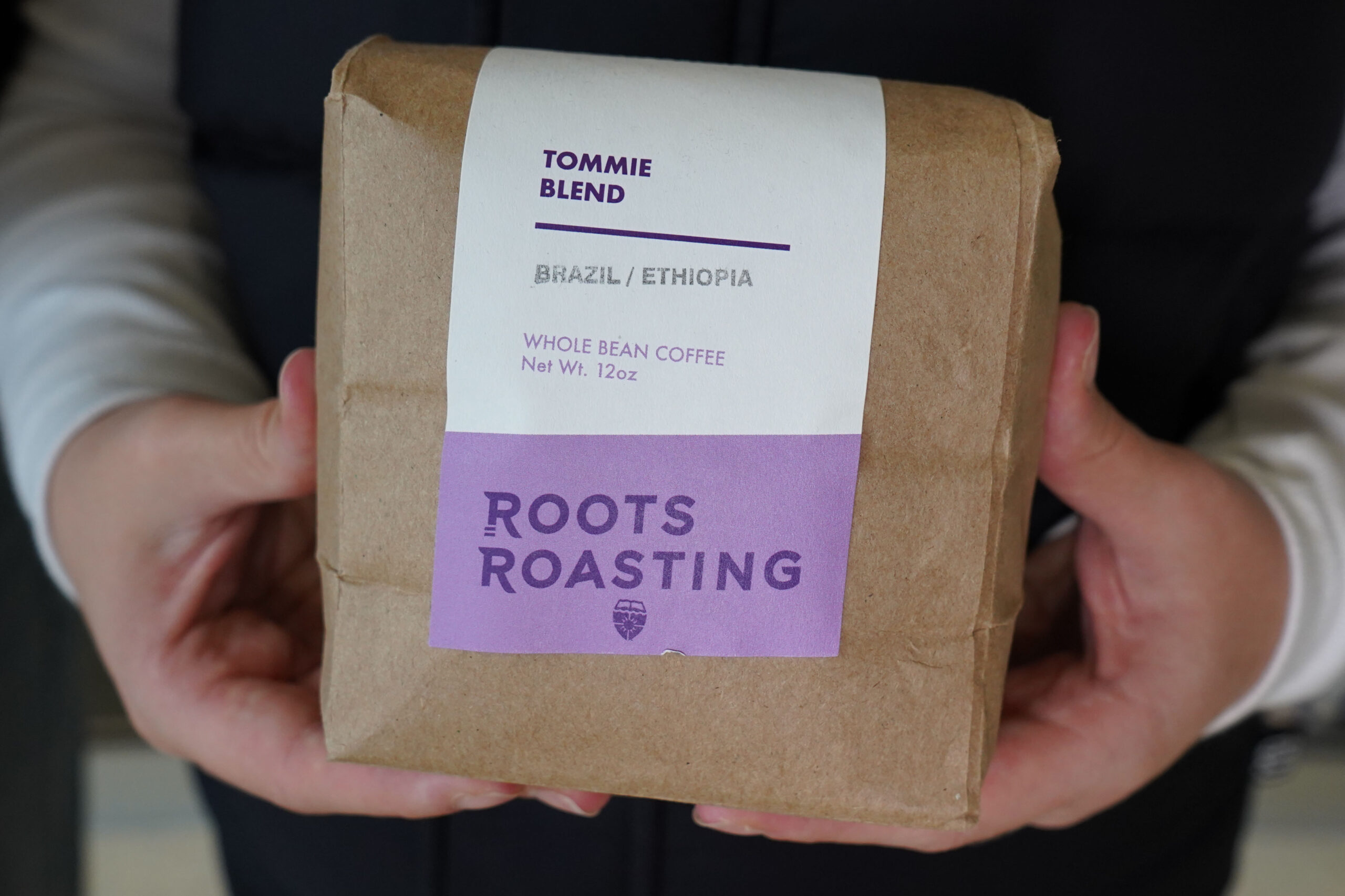 A brown bag with a white and purple paper sleeve down the middle with words on it to identify this is Tommie Blend from Roots Roasting.
