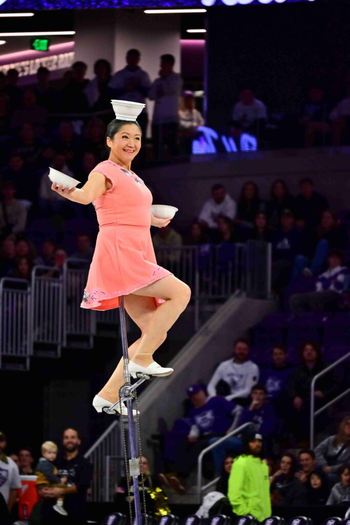 A performer balancing two bowls on her head, one in each hand while riding an eight foot tall unicycle.