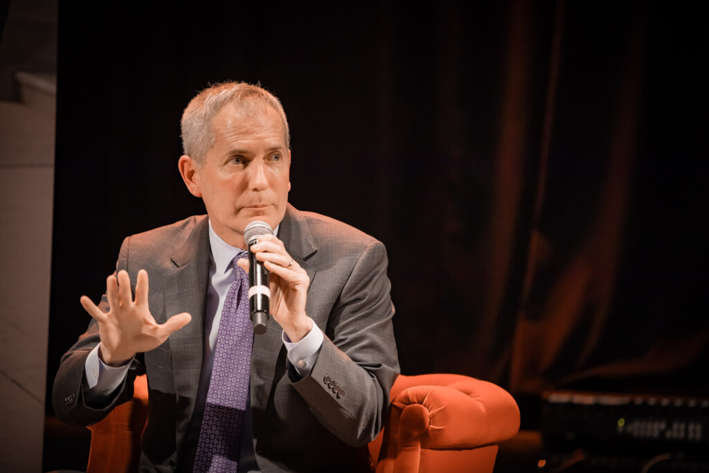 St. Thomas President Rob Vischer sits in an orange chair holding a microphone, speaking with one hand raised slightly. He is wearing a gray suit and purple tie against a dark backdrop.