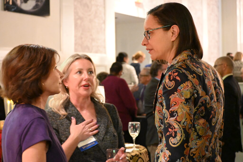 Three women stand together at a reception, smiling and talking. One holds a glass of white wine while another gestures as she speaks. People mingle in the background.