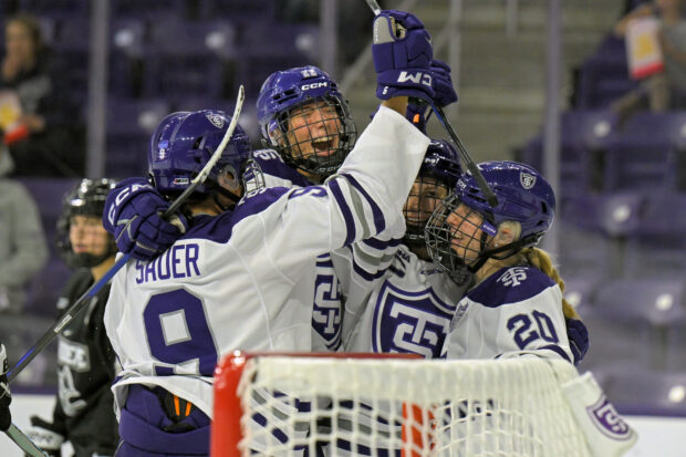 Four members of the St. Thomas women's hockey team hug in front of the net after scoring a goal during match.