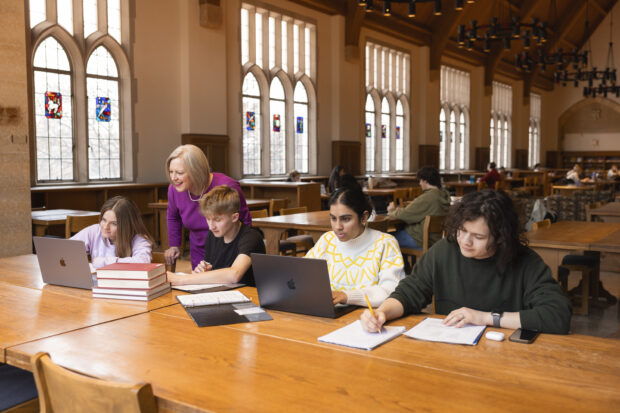 Four students sit at a table. One woman stands over a students shoulder.
