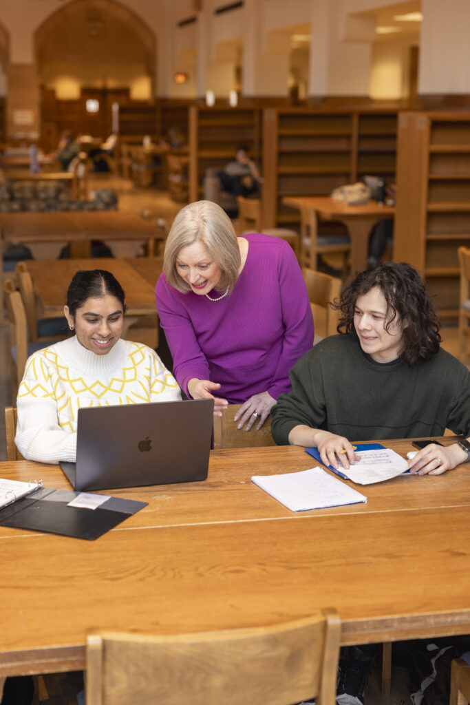 Four students sit at a table. One woman stands over a students shoulder.