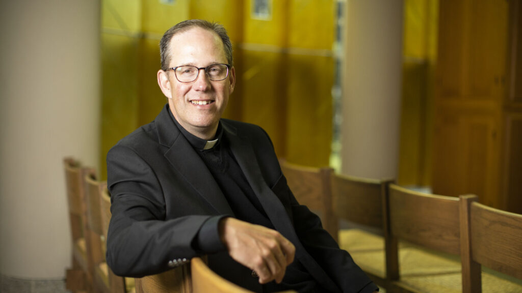 A man wearing a black clerical suit with a Roman collar and glasses sits sideways on a wooden pew, smiling at the camera. The background features warm yellow tones, wooden chairs, and architectural columns, suggesting a church or chapel setting.