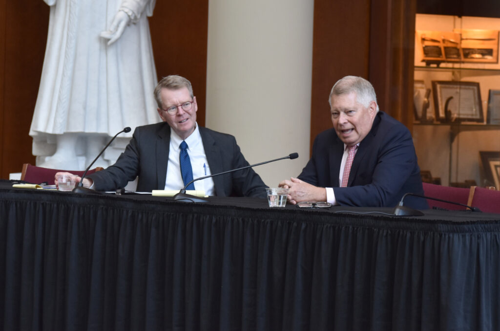 Two men dressed in suits are sitting at a panelist table.
