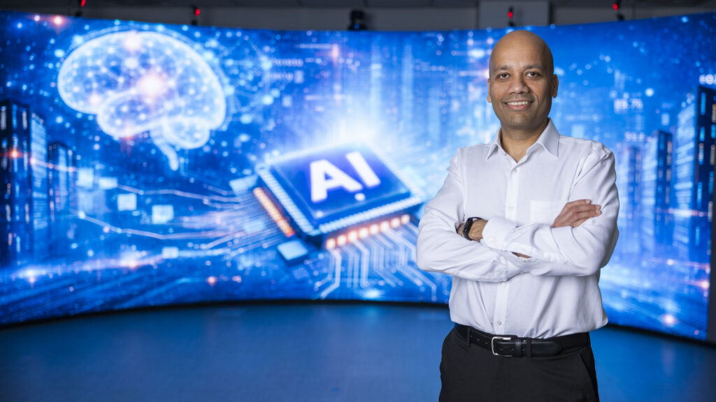 Dr. Manjeet Rege standing with arms crossed in front of a digital AI-themed background featuring a brain graphic and a computer chip labeled “AI.”