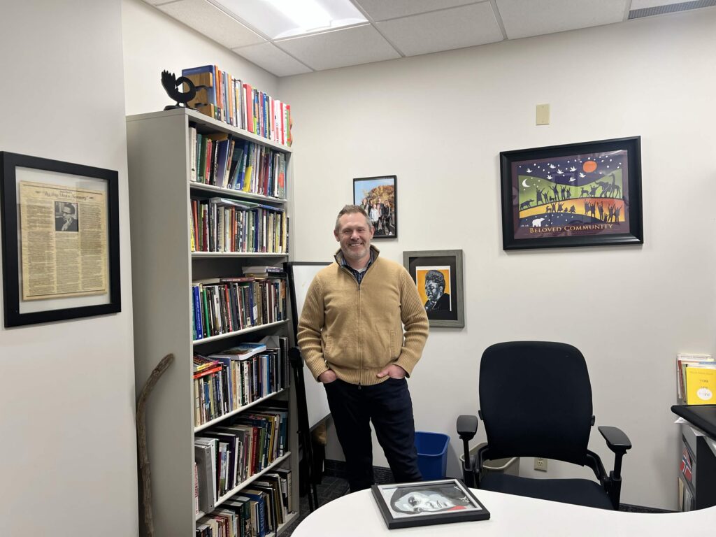 A man smiles and stands in the middle of an office with portraits and book.