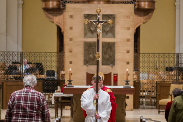 An individual walks down church aisle with a crucifix symbolizing Christ’s entry into Jerusalem while immediately foreshadowing his passion and death.