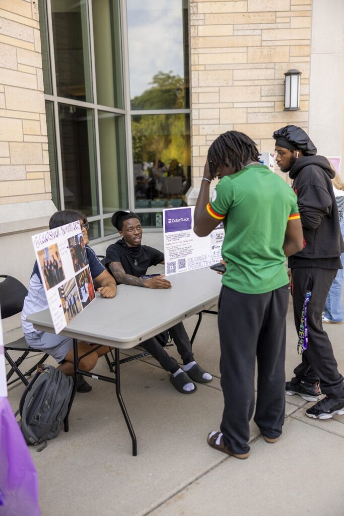 Two students representing ColorStack sit at a table talking with two students guests visiting their station at a fair.
