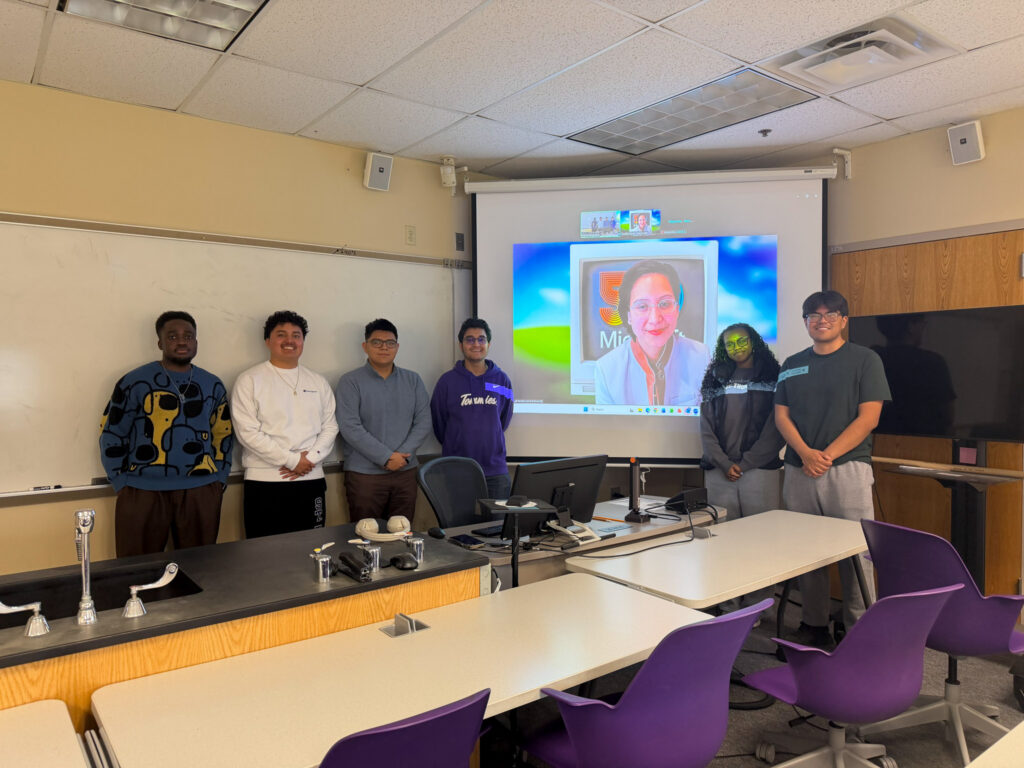 A group of students pose for a photo next to the projector showcasing their virtual speaker.