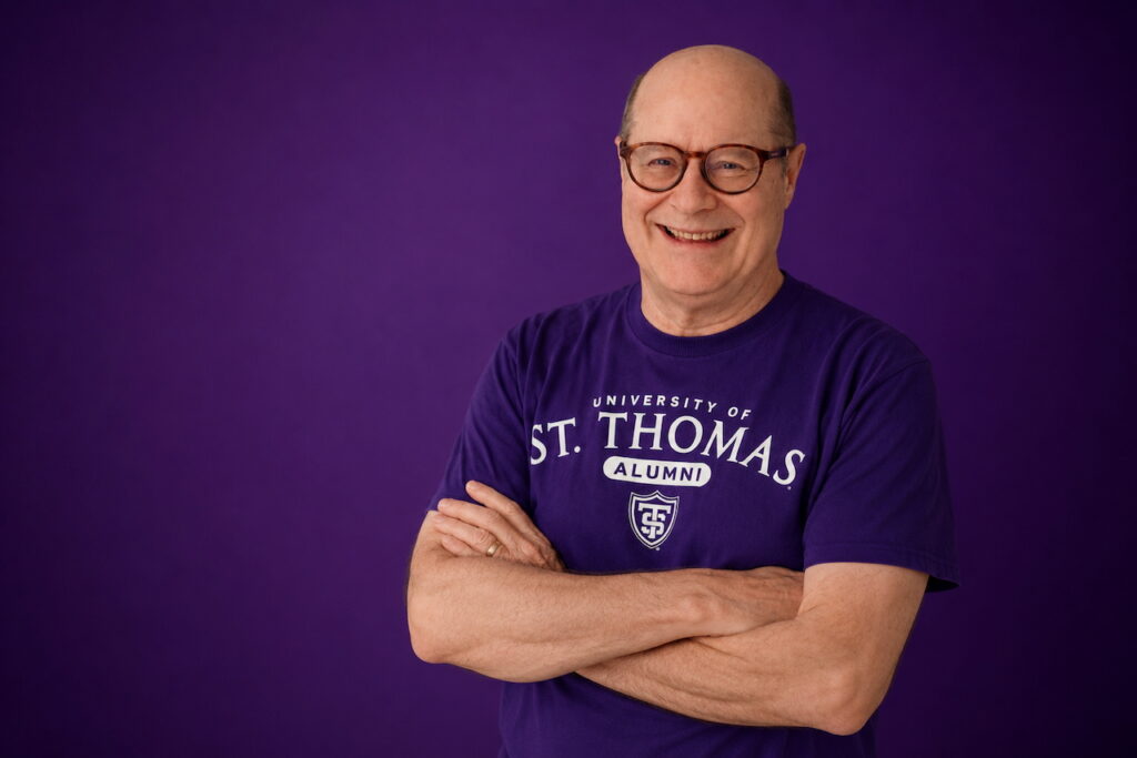 Marty Strommer crosses his arms and smiles for a photo. He wears purple with a purple background.