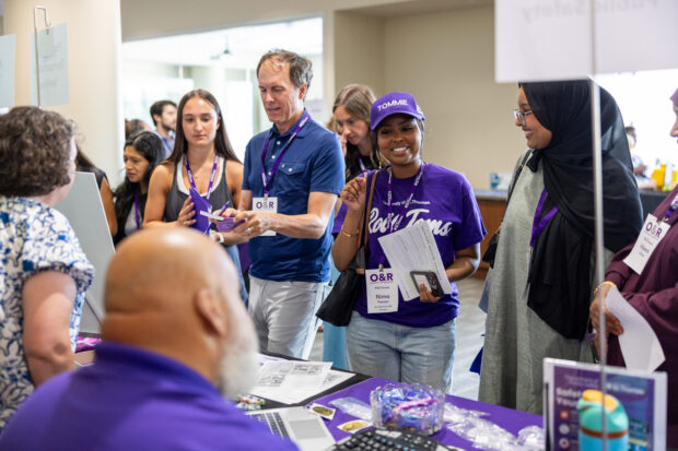 Students standing in front of a table engaging with staff members during an information fair.