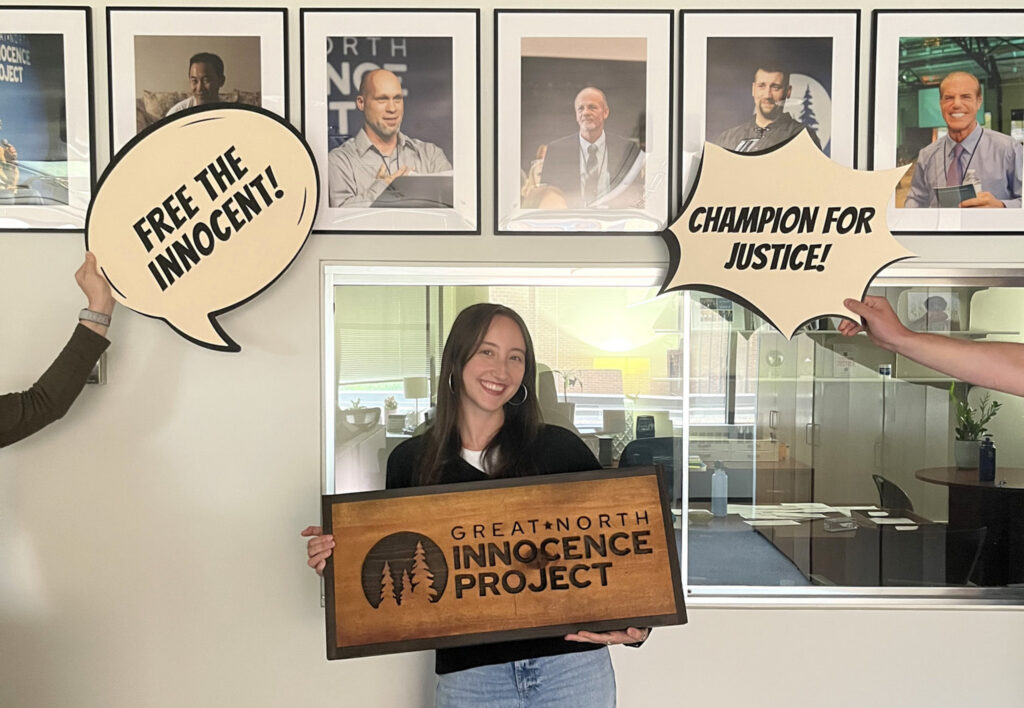 Kayla Abrams-Mahoney holds a sign that reads "Great North Innocence Project" in a hallway in front of framed pictures.