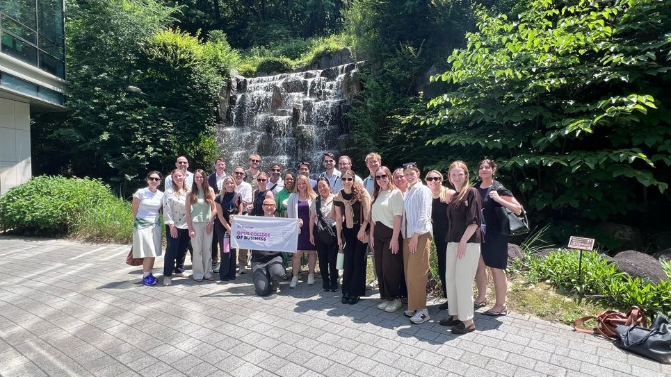 A group of over two dozen graduate students pose for a photo abroad near a waterfall.