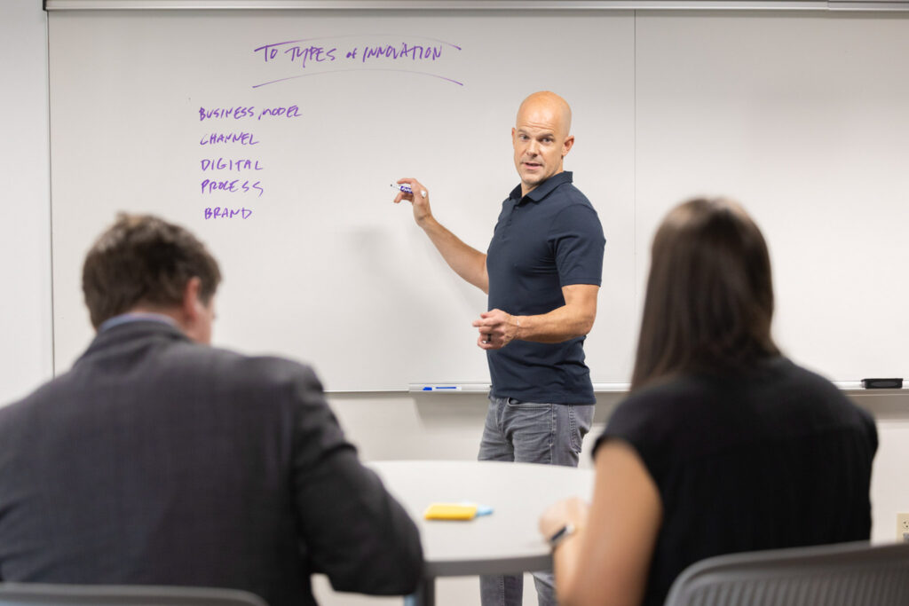 Ryan Backer stands in front of the class next to the white board with a dry erase marker in his hand.