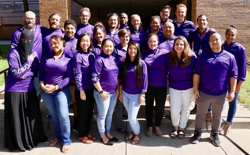 A group of 23 wear matching purple track jackets and smiles for a group photo.