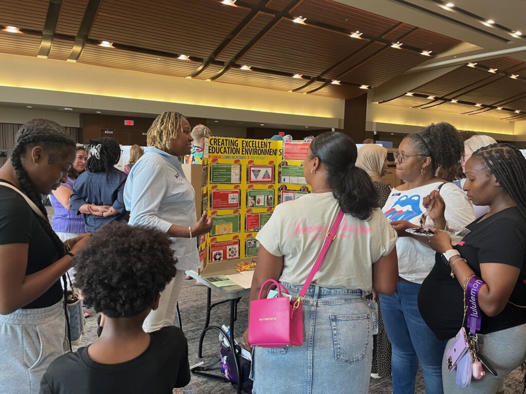A group huddles around a booth listening to a presenter at a fair.
