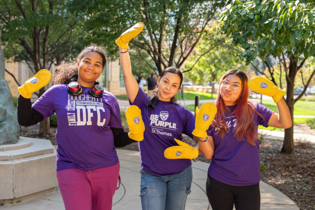 Three DFC students pose for a photo wear St. Thomas purple shirts and yellow clapper mittens.