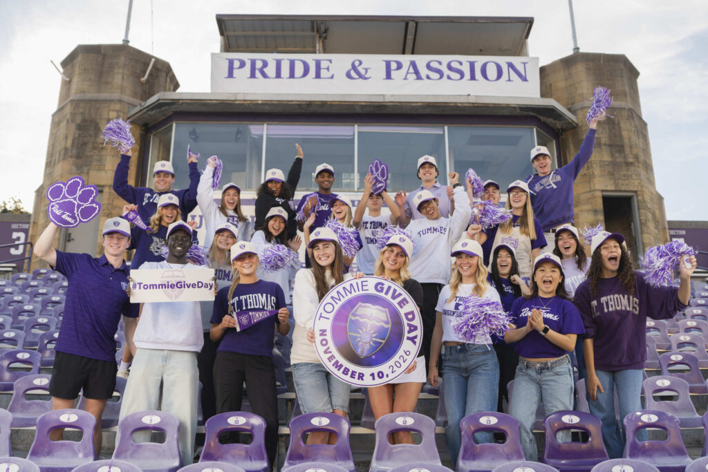 A group of students in the bleachers at O’Shaughnessy Stadium holding up a sign and posing for a Tommie Give Day campaign photo.